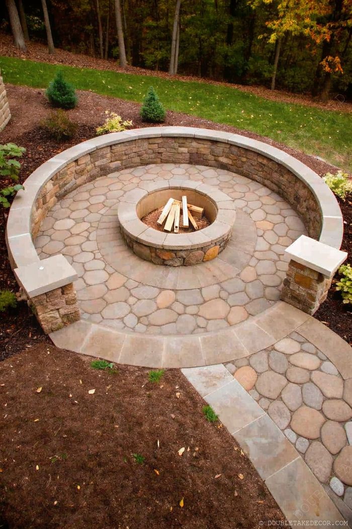 Circular stone fire pit surrounded by mosaic patterned paving, with lush greenery and evergreen shrubs creating a rustic, inviting outdoor space.