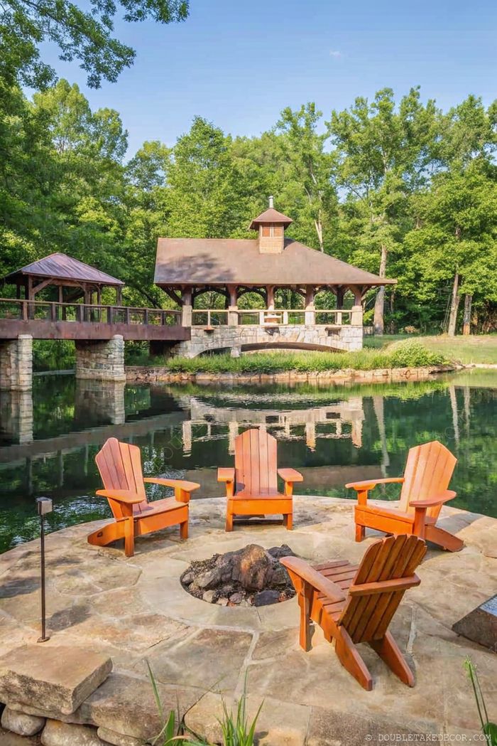 Adirondack chairs surround a stone fire pit near water, with a bridge leading to a charming gazebo amid lush greenery