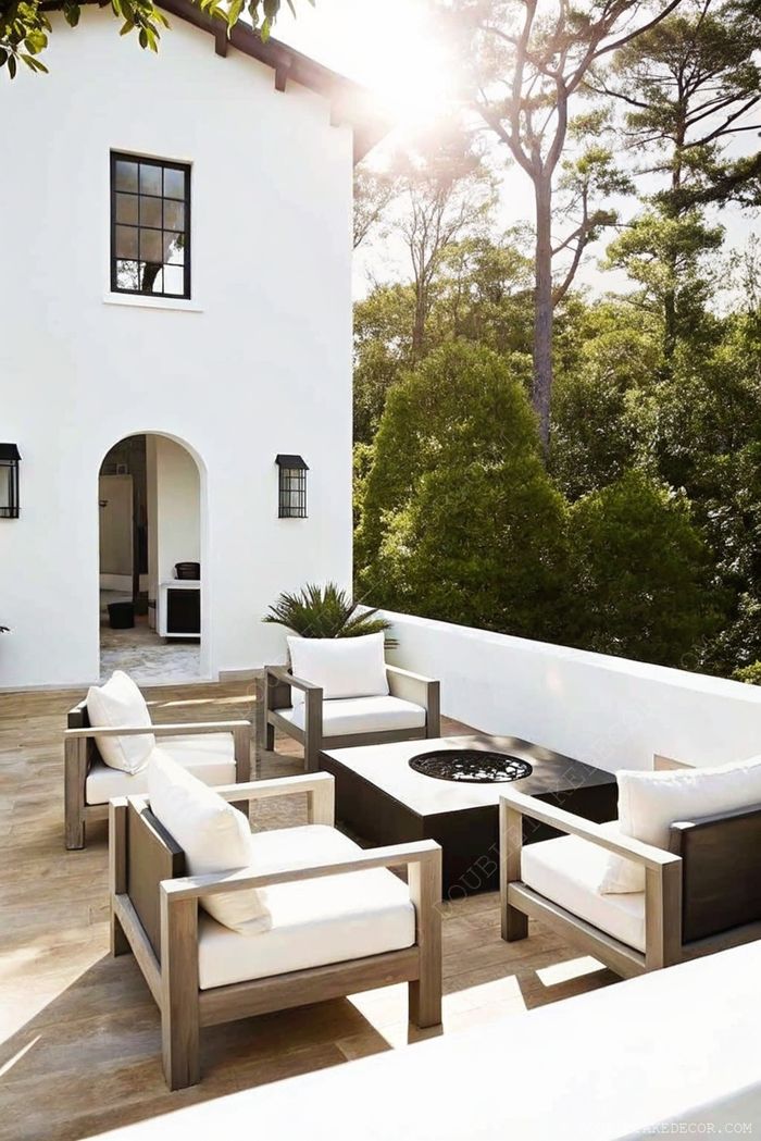 Serene outdoor area with wooden armchairs, black fire pit table, and white-washed walls surrounded by lush greenery