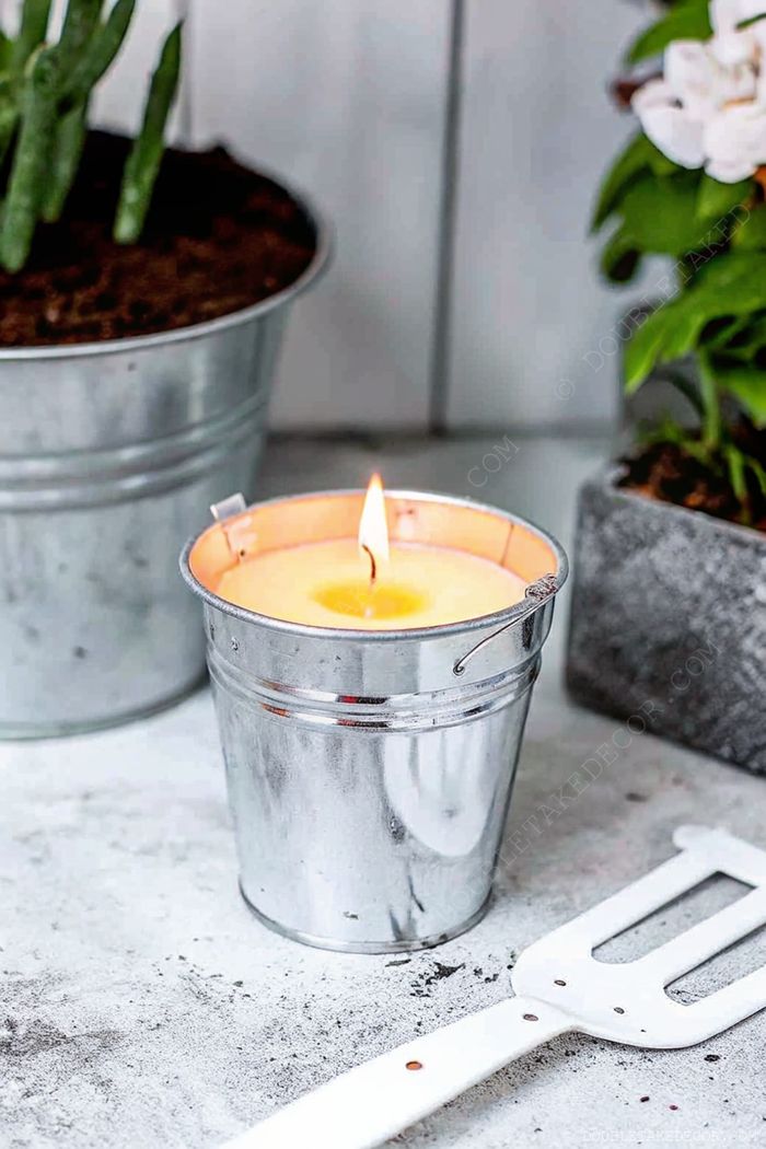 Galvanized metal bucket holding a warm yellow candle surrounded by potted plants and a gardening tool, creating a cozy outdoor atmosphere