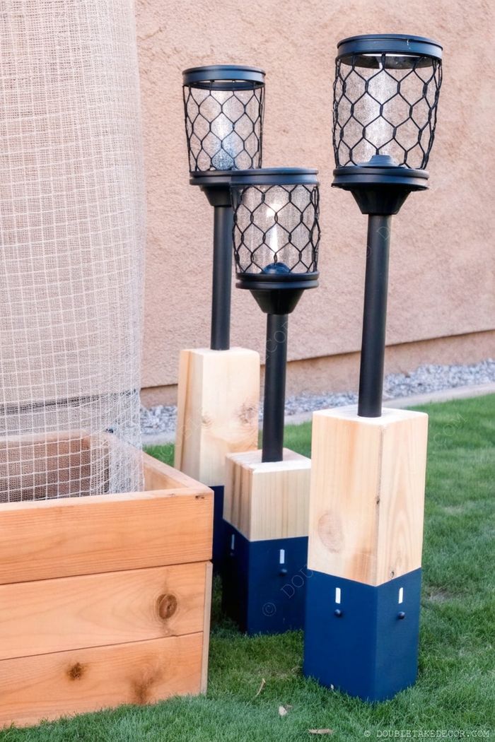 Three outdoor LED garden lights with glass lanterns, wire mesh design, and wood-to-navy blue bases standing on green grass near a wooden planter.