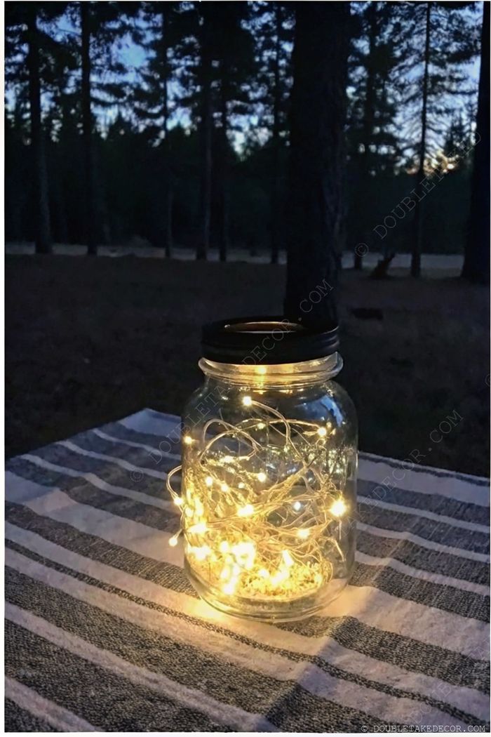 Glass jar filled with twinkling fairy lights on a striped cloth, creating a warm glow against a backdrop of trees at dusk