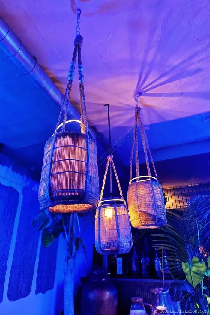Stylish hanging woven lanterns with braided cords casting warm light against a blue background, complemented by greenery and glass bottles.