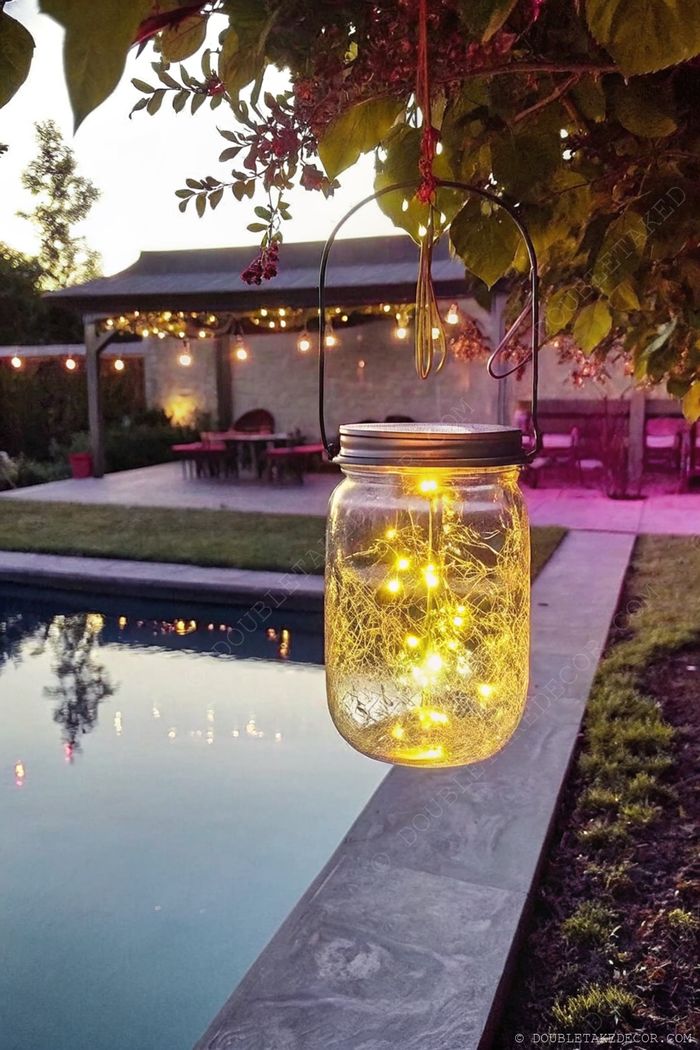 Mason jar with fairy lights hangs above a pool at dusk, illuminating the water with string lights and red chairs in background.