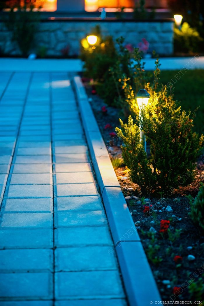 Softly lit outdoor pathway with square pavers, flanked by shrubs and flowers, illuminated by warm glowing lanterns