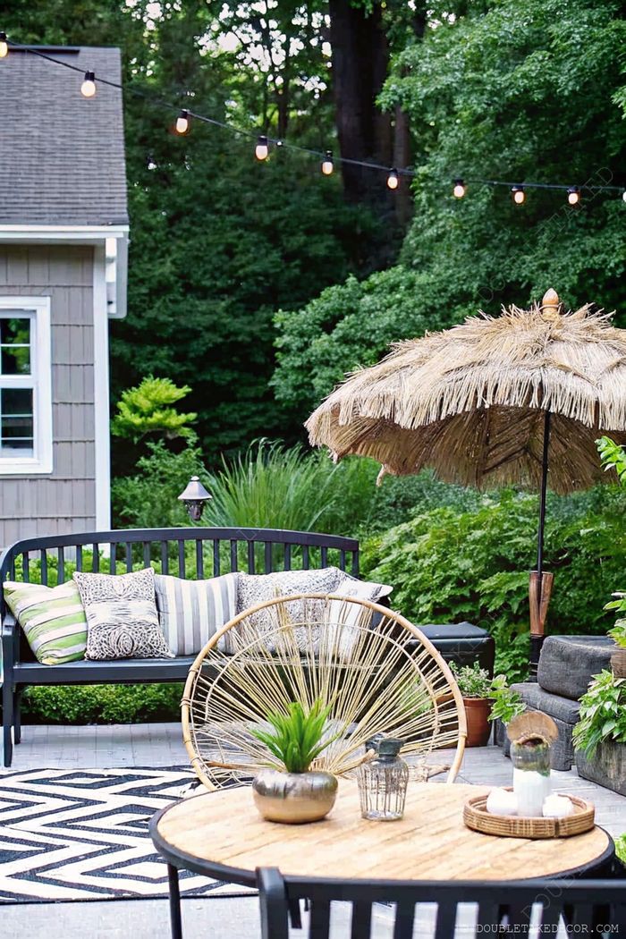 Espace extérieur cosy avec banc noir, fauteuil rond en rotin, guirlandes lumineuses, parasol en paille et éléments déco sur un tapis à motifs.