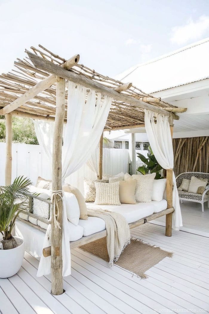 Espace détente extérieur paisible avec pergola en bois, rideaux blancs et lit de jour confortable avec coussins et plaid neutres.
