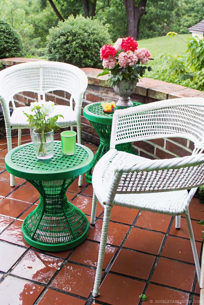 Coin salon extérieur cosy avec chaises blanches tressées autour d'une table verte sablier décorée de roses, de rafraîchissements et de carreaux mouillés par la pluie.