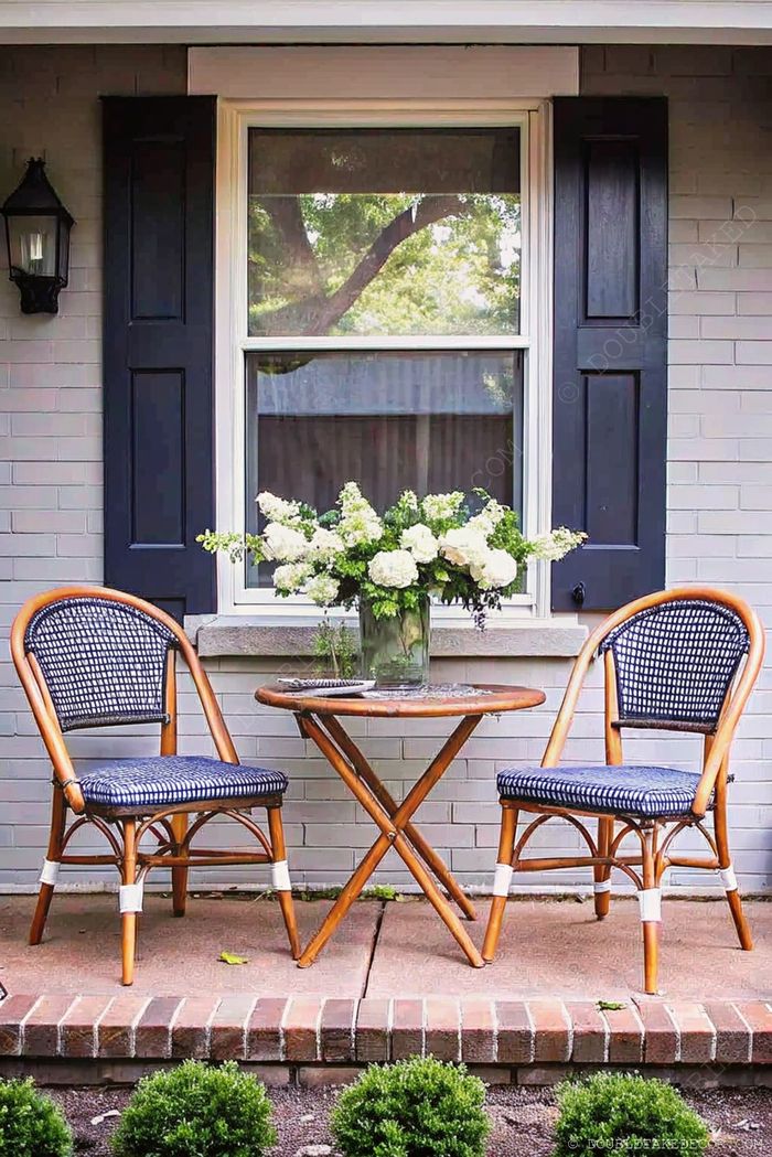 Table ronde en bois avec fleurs au centre entre deux fauteuils en osier avec coussins bleus, adossés à un mur en briques avec volets.
