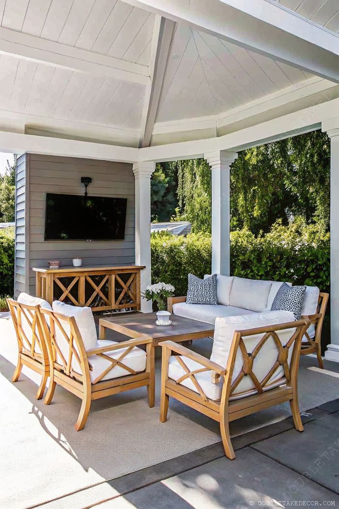 Espace extérieur élégant avec canapé blanc, fauteuils et table basse en bois sous une terrasse couverte, TV fixée au mur et verdure naturelle.