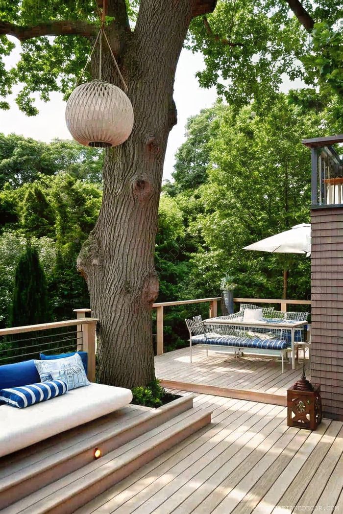 Terrasse extérieure paisible avec banquettes et coussins sous un arbre équipé d’une suspension lumineuse, et chaises de repas métalliques sous un parasol.