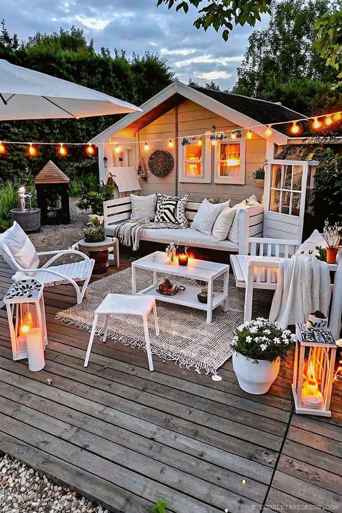 Coin salon extérieur douillet avec canapé en bois blanc, coussins et lanternes devant un charmant cottage éclairé.