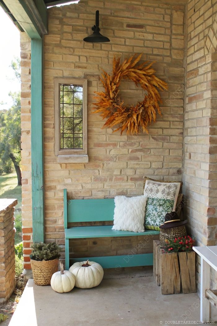 Cozy outdoor entryway with brick walls, autumn wreath, teal bench with cushions, and seasonal white pumpkins and plants.