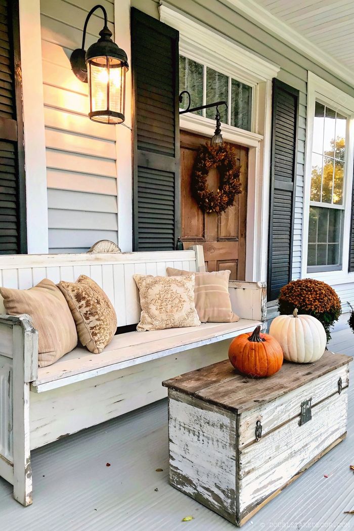 Charming front porch with white bench, pumpkins on wooden chest, black shutters, and fall wreath creating a cozy autumn atmosphere.