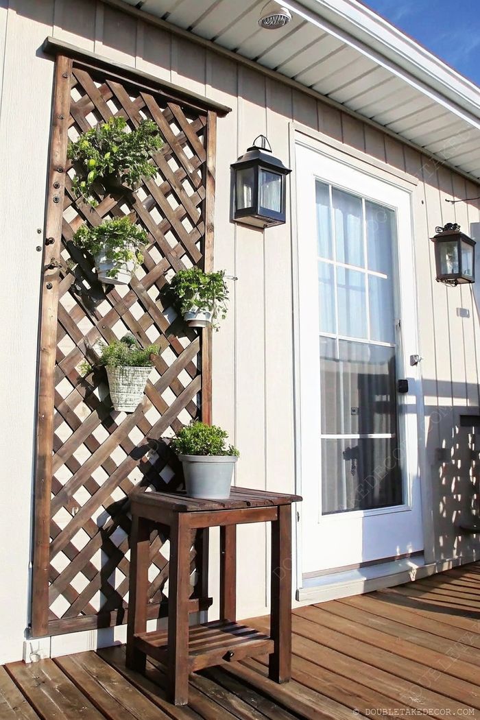 Charming outdoor space featuring wooden lattice trellis with potted plants, white door with lanterns, and wooden side table on deck.