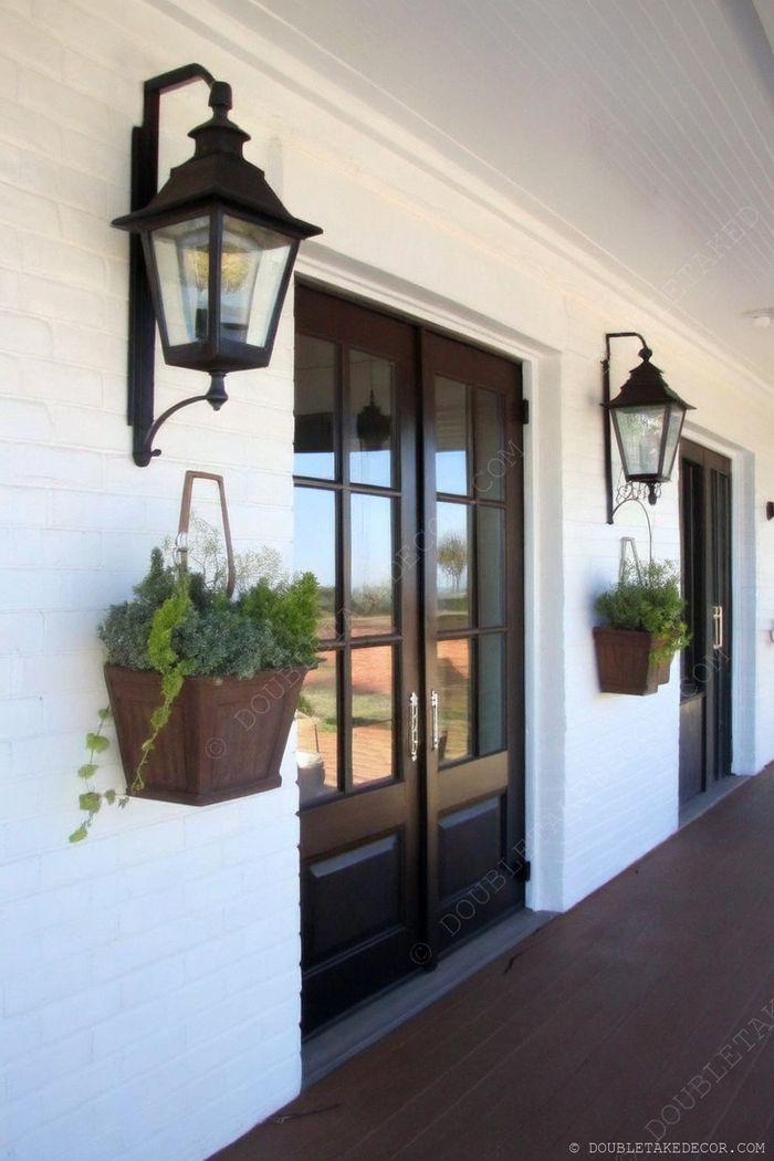 Elegant entryway featuring dark wooden doors with vintage lantern sconces and wooden planters with greenery against a white brick backdrop.