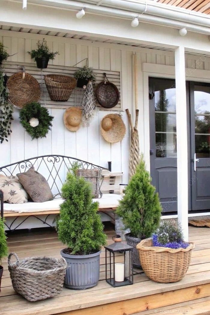 Inviting white-paneled porch featuring woven baskets, wreath, wrought iron bench with pillows, and potted plants for a serene outdoor retreat.