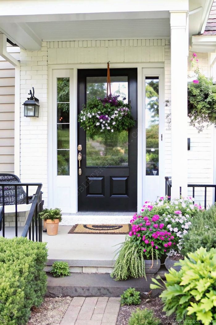 Inviting front porch featuring a striking black door against white brick, adorned with hanging purple flowers and flanked by comfortable seating.
