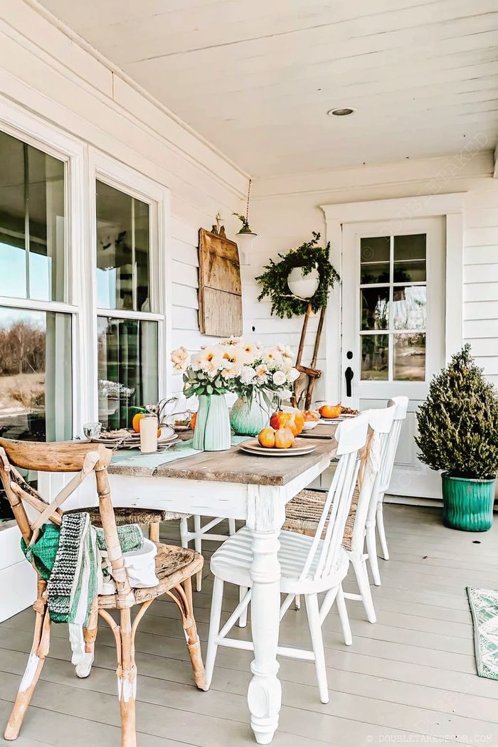 A rustic wooden dining table with mixed chairs on a bright white porch, decorated with fresh flowers and citrus accents for casual outdoor meals.