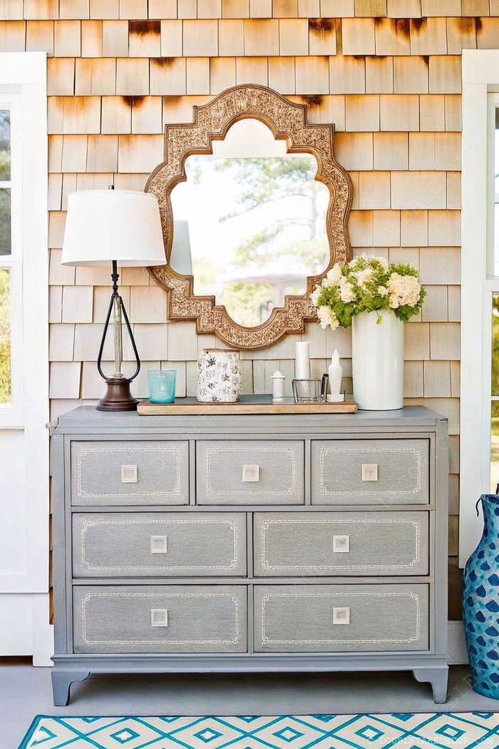Stylish gray dresser with mirror on wooden shingle porch wall, decorated with lamp, turquoise vase, candles and fresh flowers