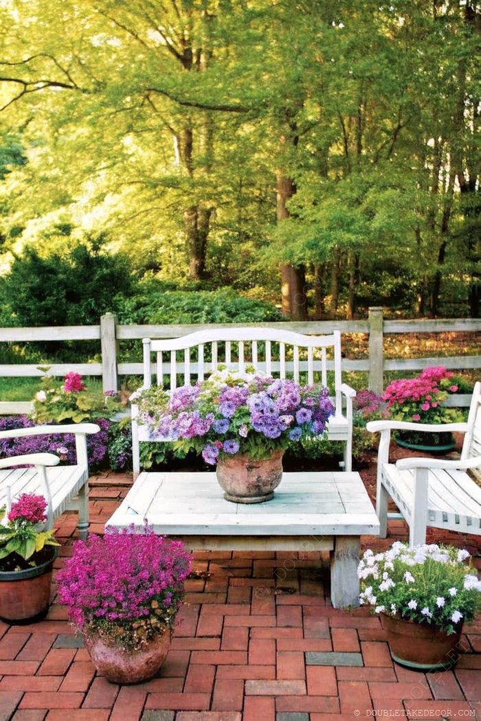 Charming outdoor seating area with rustic table and white benches surrounded by colorful potted plants on brick flooring.