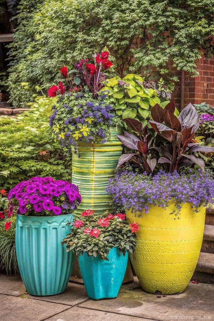 Colorful garden display with five large pots in turquoise, yellow, green, and purple, filled with petunias and mixed blooms against lush greenery.