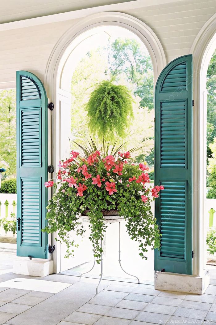 Elegant indoor space with turquoise shutters, arched entrance, and overflowing planter of pink flowers and cascading foliage on stone flooring.