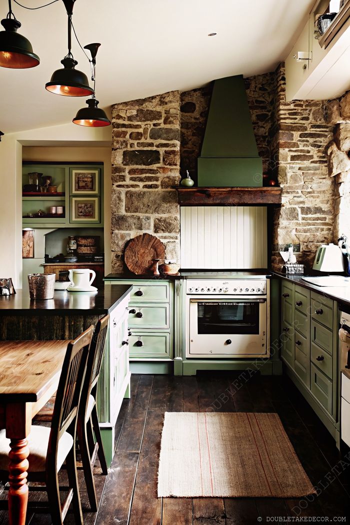 Stone-walled kitchen with soft green cabinets, dark countertops, and warm wooden dining furniture creating a cozy, stylish space.