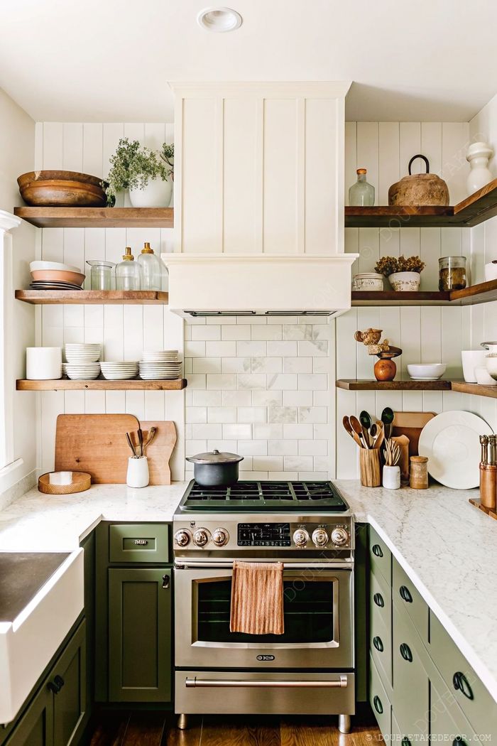 Modern kitchen featuring rich green cabinetry, marble countertops, and wooden open shelving with decorative items above a stainless steel stove.