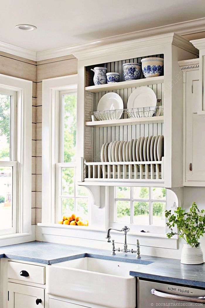 White open shelving with blue and white ceramics, farmhouse sink, dark countertops, and natural light creating a warm kitchen atmosphere.