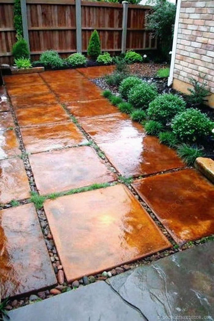 Earthy concrete stepping stones form a garden pathway, surrounded by pebbles, grass, and trimmed shrubs with a wooden fence backdrop.