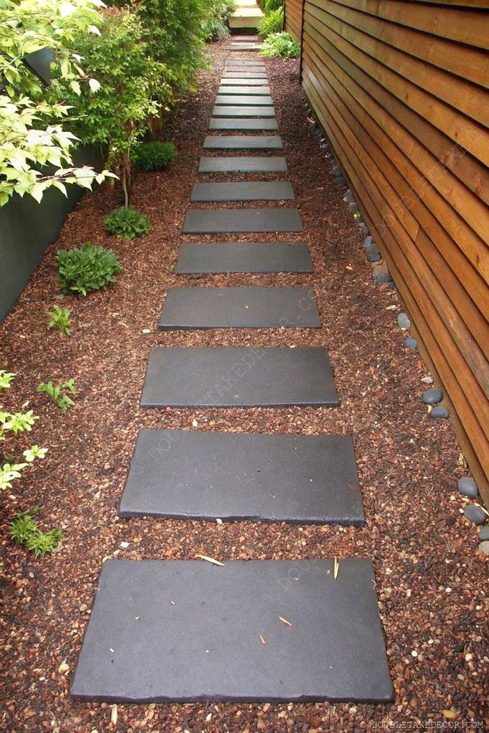 Rectangular black stepping stones create a linear garden path, bordered by lush greenery and smooth pebbles on mulched earth.