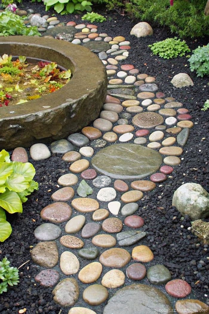 Colorful pebble garden pathway winding towards a circular stone water feature, surrounded by lush greenery and foliage.
