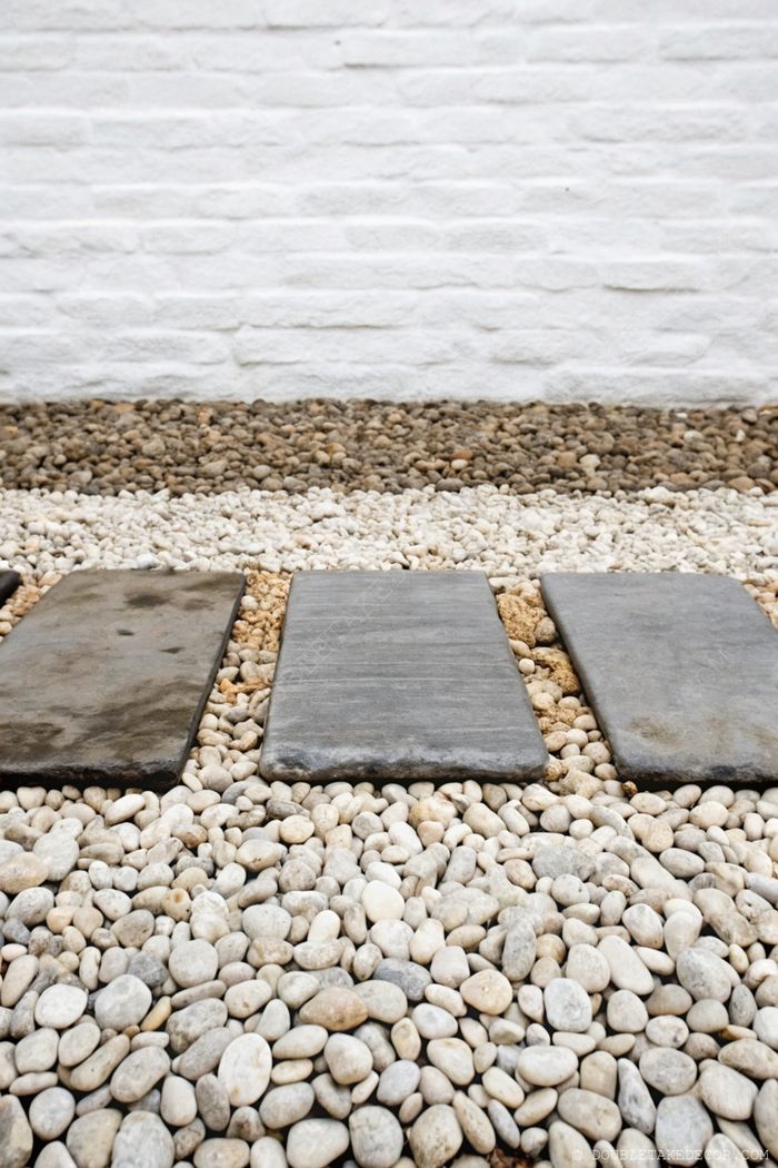 Rectangular stone slabs creating a minimalist garden path over mixed brown and white pebbles against a textured white wall.