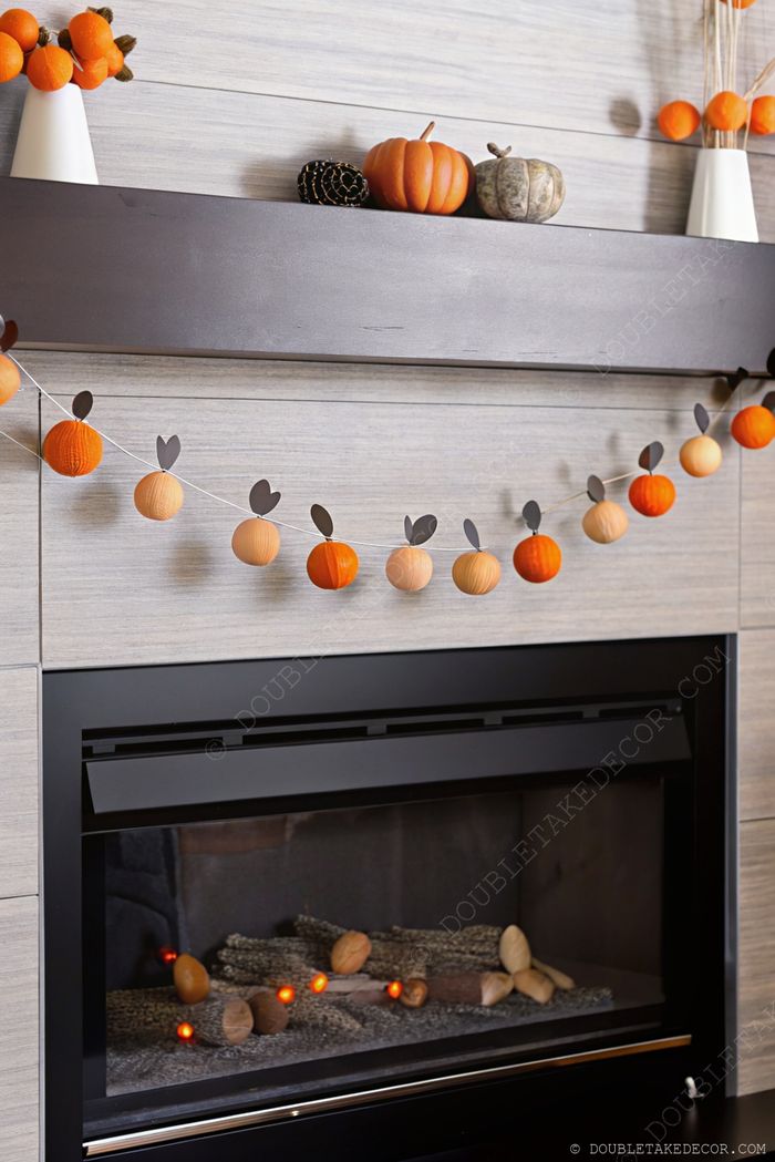 Autumn fireplace decor featuring a wooden mantel with colorful pumpkins and a charming pumpkin garland with black leaves above a glowing hearth.