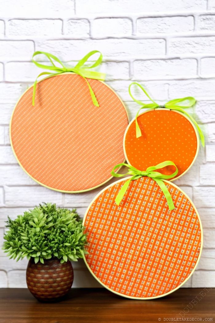 Three orange fabric circles resembling pumpkins with different patterns, hanging above a potted plant against a white brick wall.