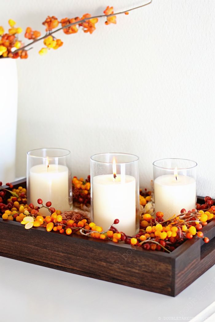 Wooden tray centerpiece with yellow-orange berries, three white candles in glass holders, and orange floral branches creating warm fall ambiance.