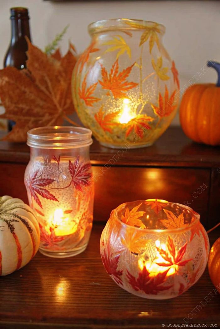 Glass lanterns adorned with autumn leaves create a warm, seasonal display alongside decorative pumpkins on a rustic wooden surface.