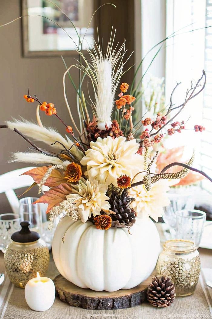 A white pumpkin centerpiece adorned with dried flowers, pine cones, autumn leaves and orange berries, displayed on a wooden slice.