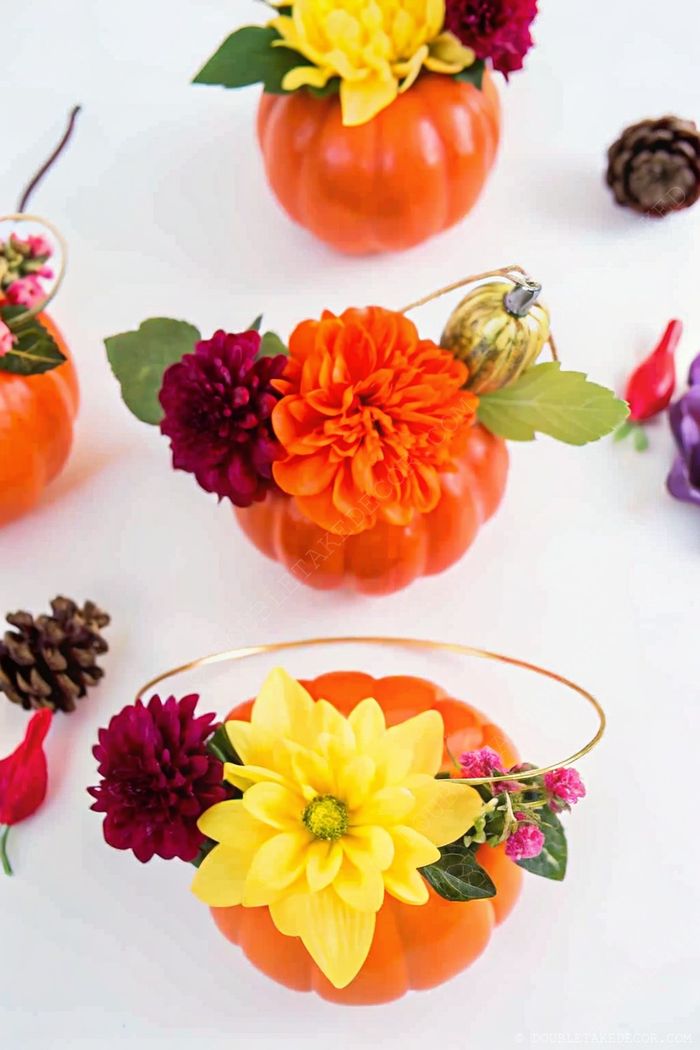 Charming fall centerpiece with small orange pumpkins transformed into vases filled with vibrant faux flowers, surrounded by decorative gourds and pine cones.