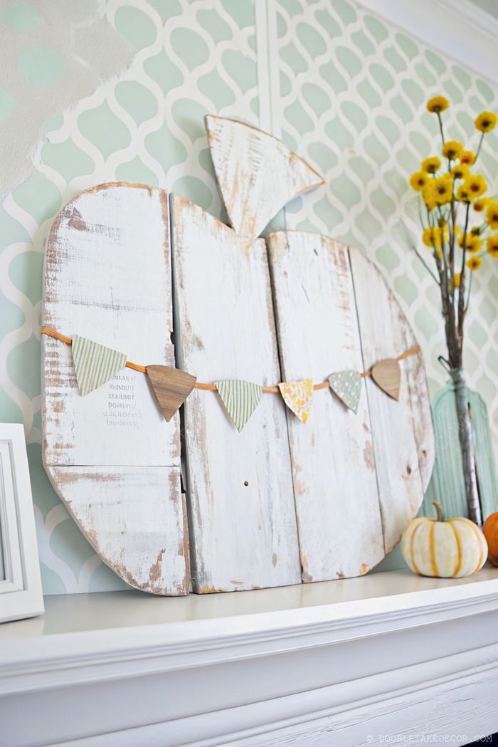 Weathered white wooden pumpkin decorated with colorful triangular flags, displayed with yellow flowers and small decorative pumpkins for fall decor.