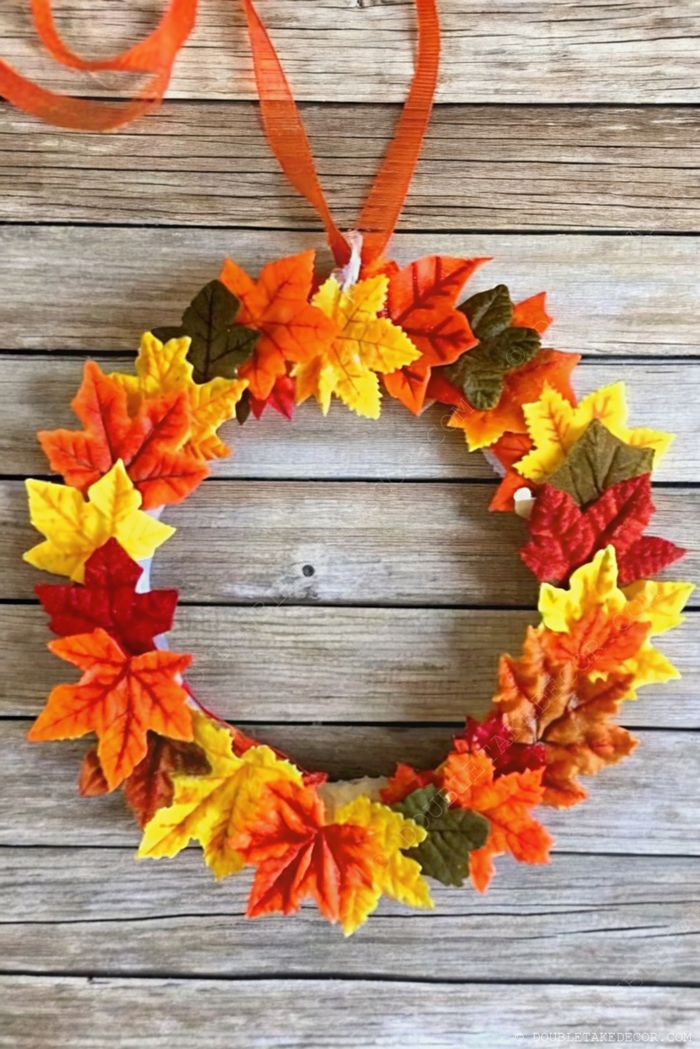 Colorful fall wreath with orange, yellow and red artificial leaves tied with sheer orange ribbon against rustic wood background.