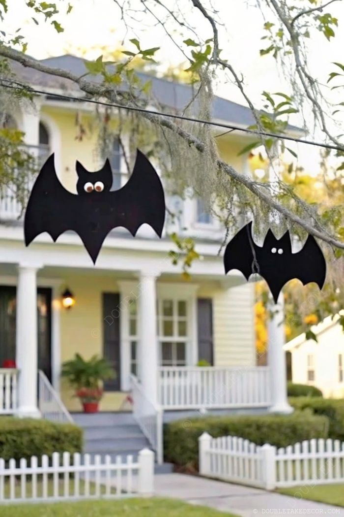 Yellow house with front porch displaying Halloween decorations - black paper bats with googly eyes hanging from tree branches.