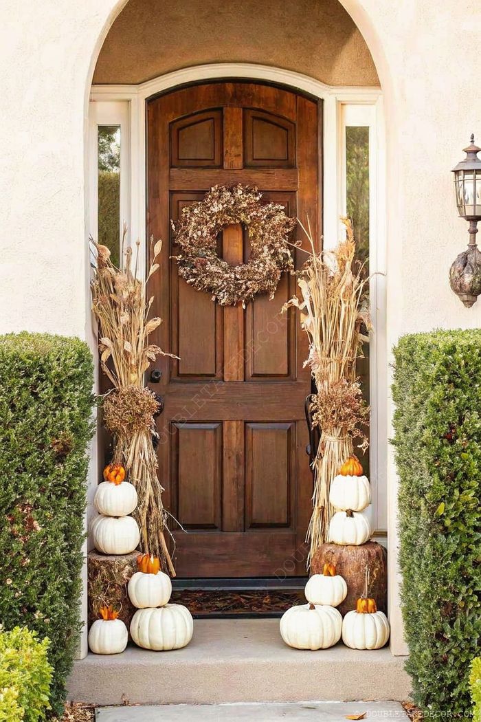 Fall front entryway with wooden door, dried wreath, white pumpkins on hay stacks, and tall grass arrangements creating harvest ambiance.
