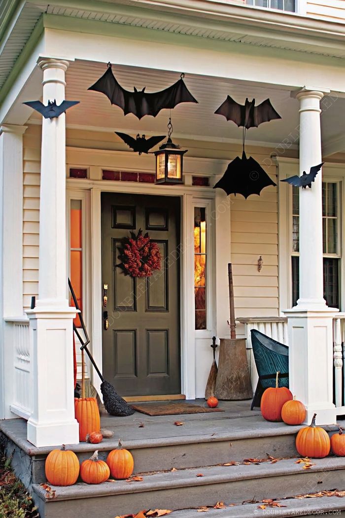 Cozy Halloween porch with green door, autumn wreath, hanging bats, orange pumpkins, and a broomstick creating a festive seasonal welcome.