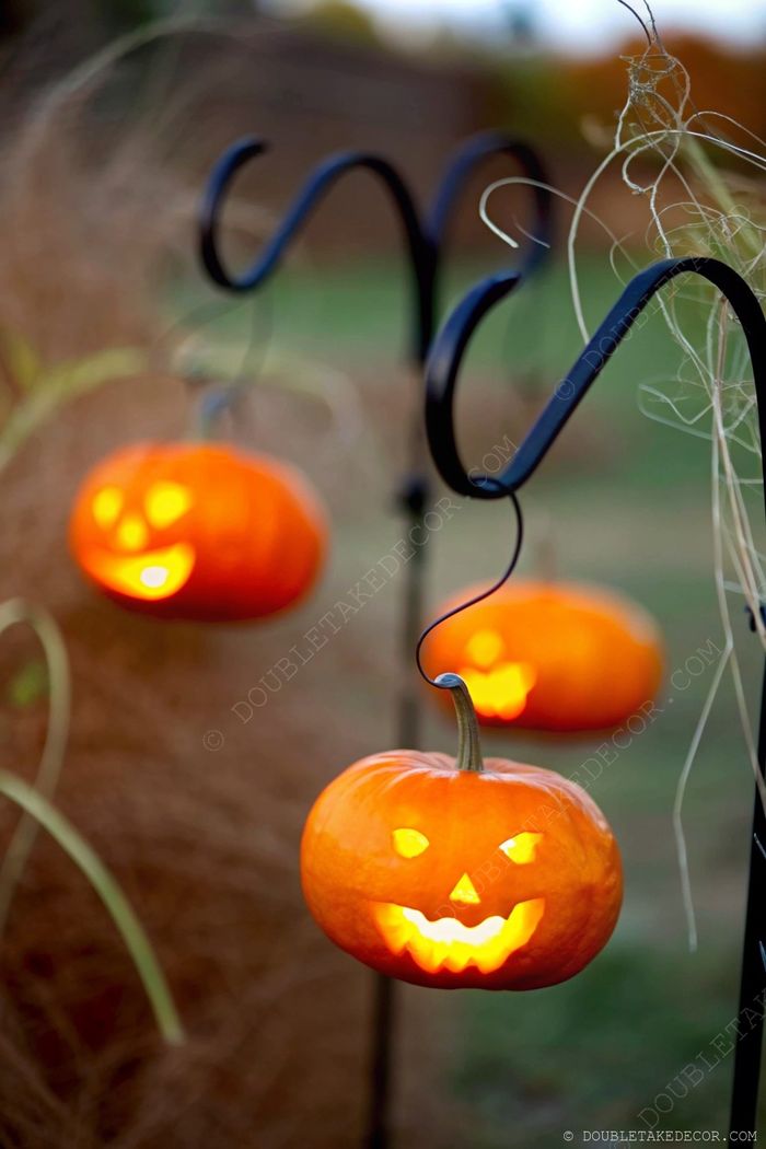 Three illuminated jack-o'-lanterns with carved faces hanging from black hooks against a rustic autumn backdrop with dry grasses