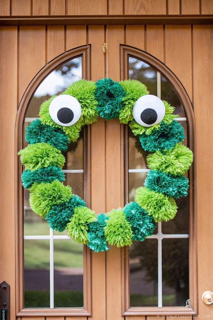 Playful Halloween wreath made from fluffy green pom-poms with large googly eyes, hanging on a wooden door as whimsical entrance decor.