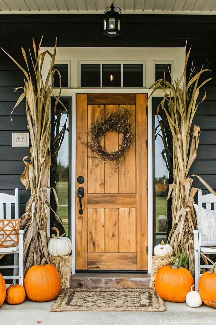 Charming front porch decorated with corn stalks, pumpkins, hay bales, and a twig wreath on wooden door, complete with white rocking chair.