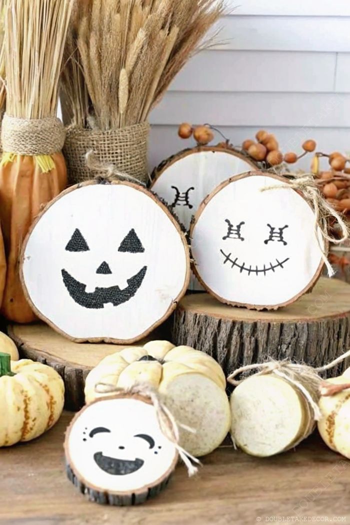 Rustic wooden slices with hand-painted faces arranged among light-colored pumpkins and dried wheat with burlap accents for fall decor.