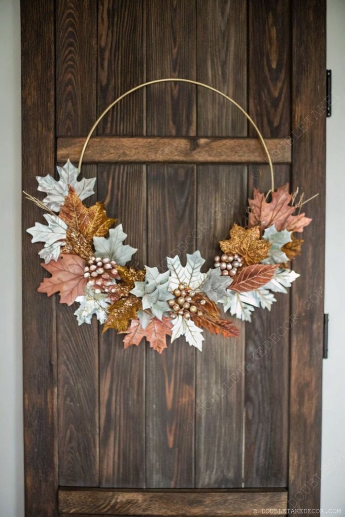 Stylish autumn wreath with orange, green and gold faux leaves arranged on a brass hoop with decorative berries, displayed on a rustic wooden door.